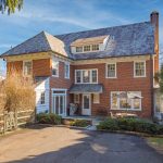 Two-story red brick house with a slate roof, small patio with umbrella and table, driveway and a portable basketball hoop