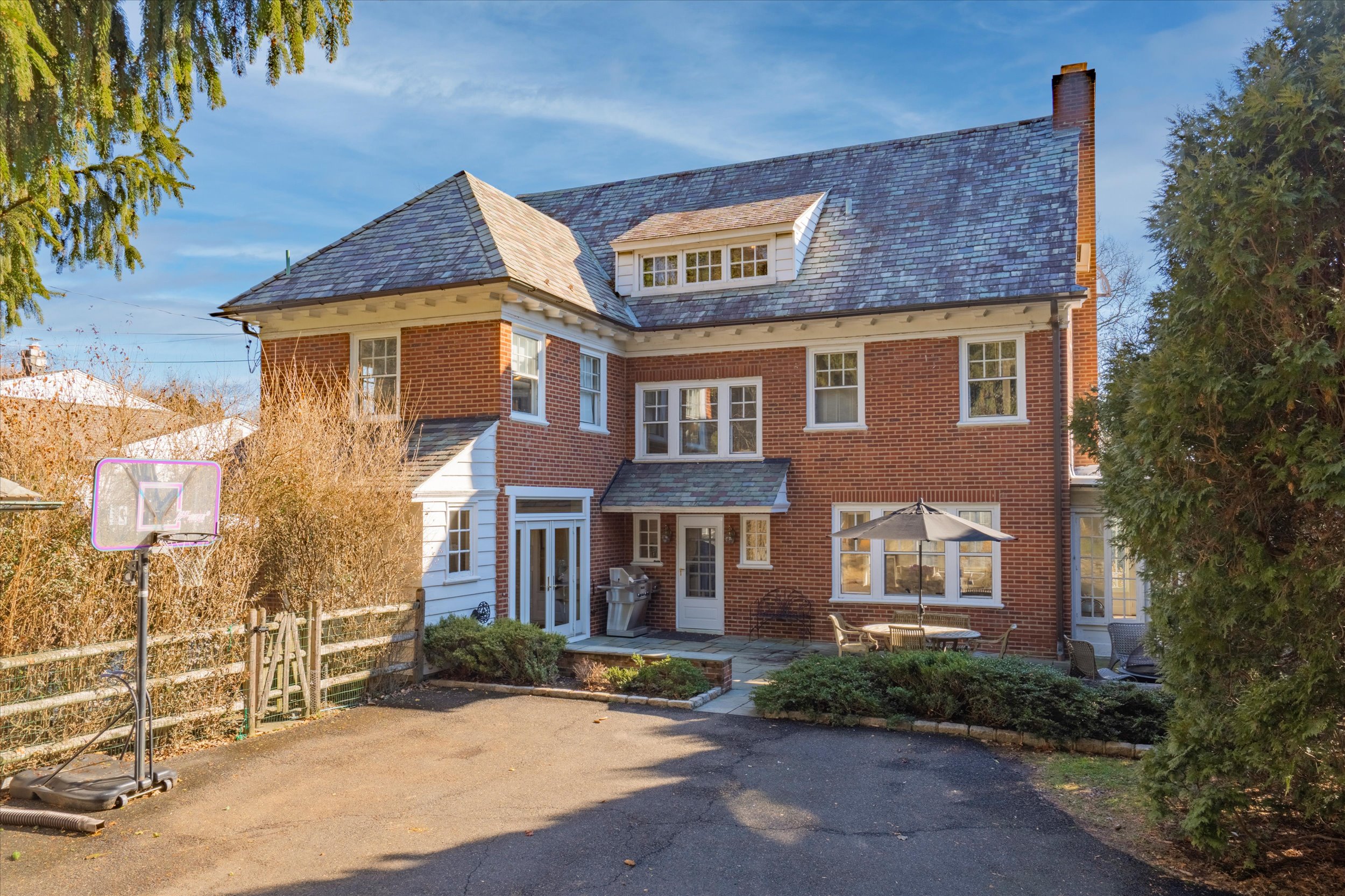 Two-story red brick house with a slate roof, small patio with umbrella and table, driveway and a portable basketball hoop