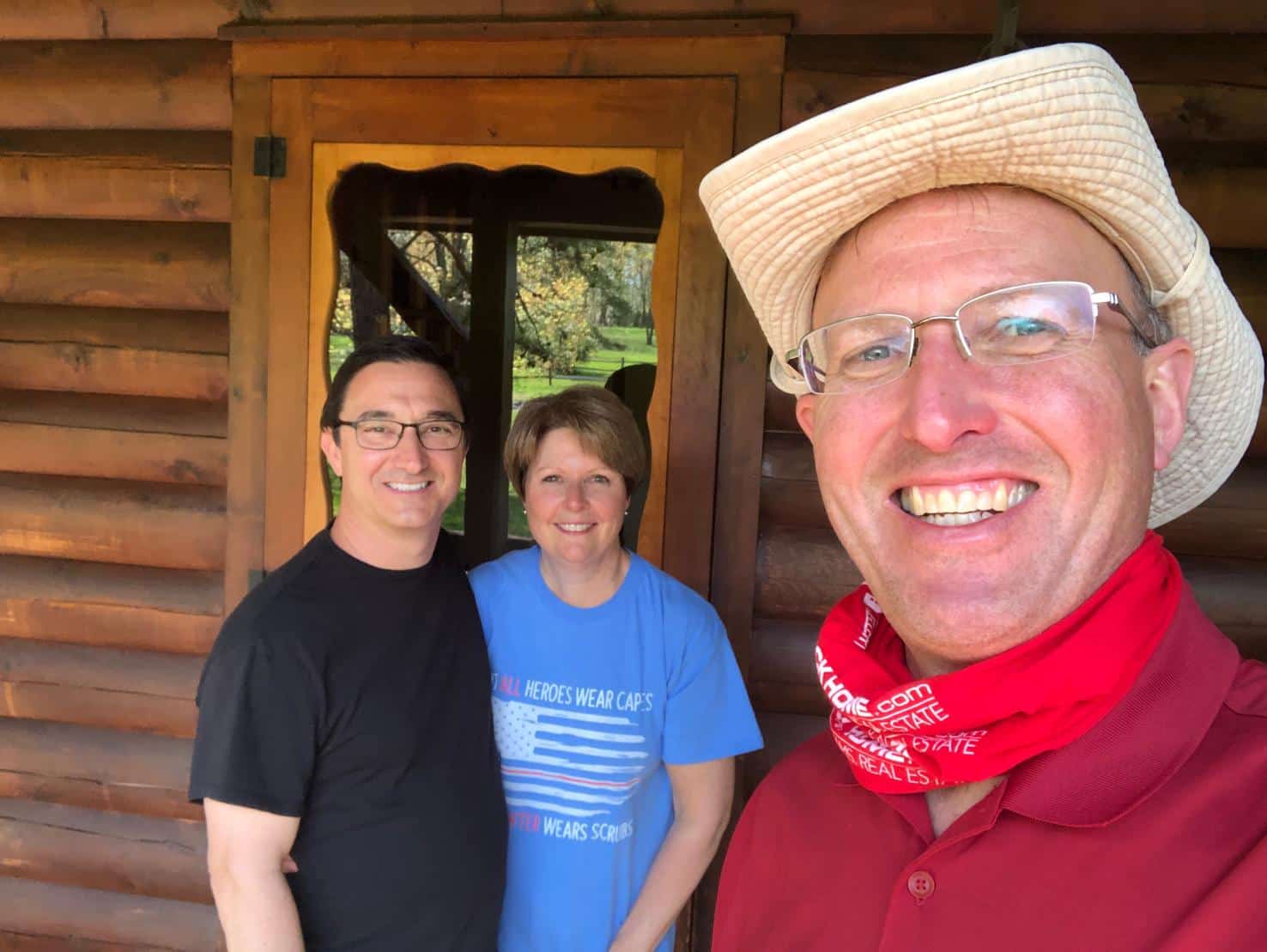 Smiling man in a straw hat and red bandana takes a selfie with a smiling couple standing in front of a wooden cabin.