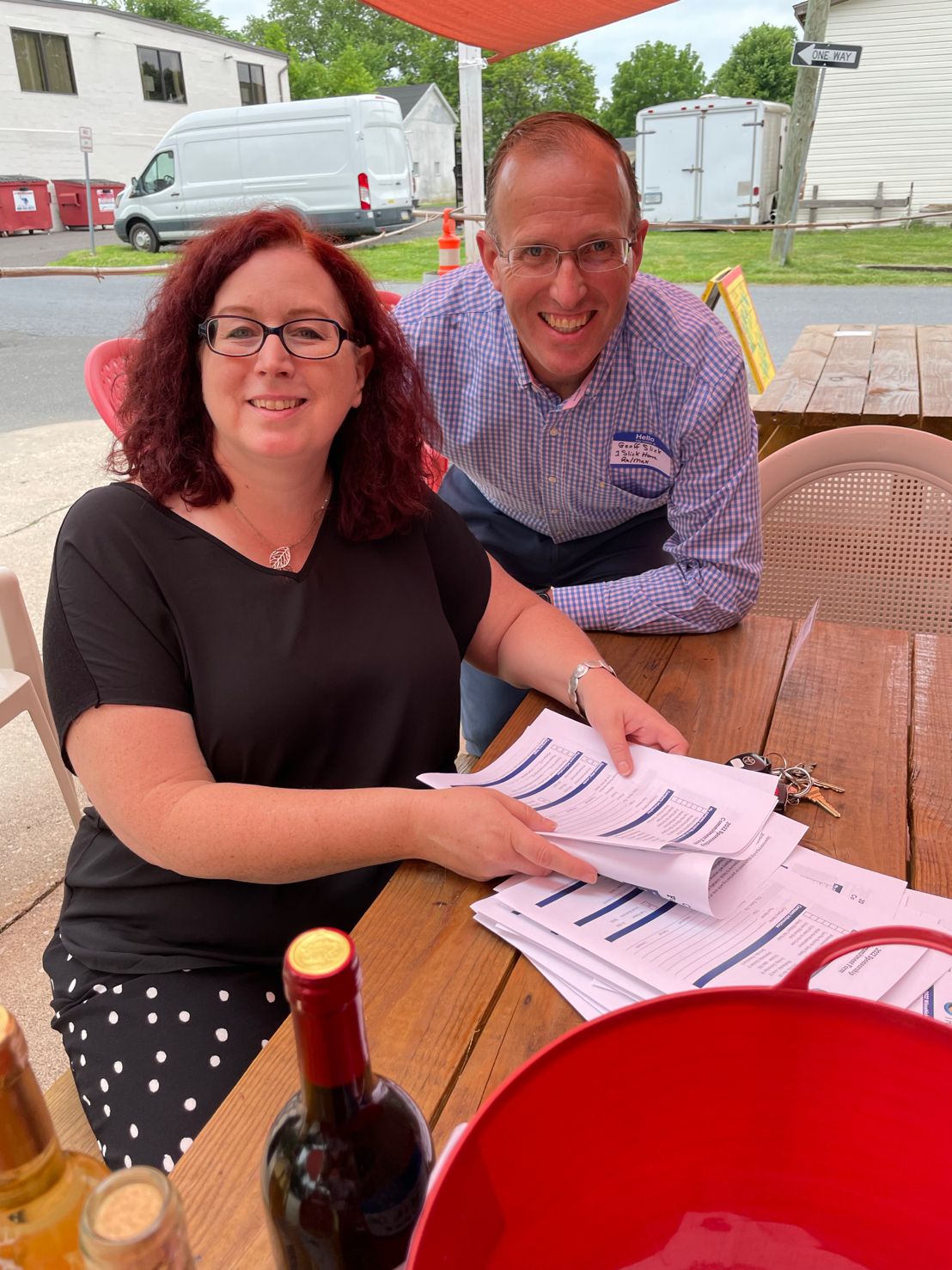 Woman with glasses and red hair reviews printed forms at a picnic table while a smiling man in a checked shirt leans in behind her.