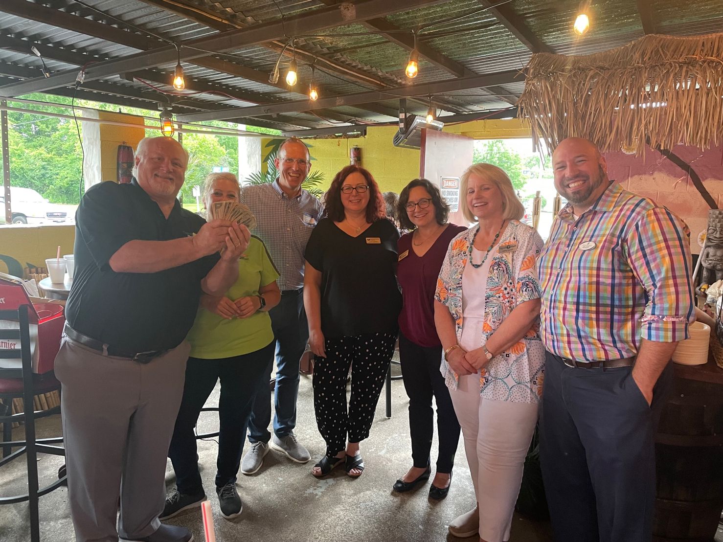 Man takes a selfie with a smiling group of seven adults posing inside a community hall with round tables and folding chairs.