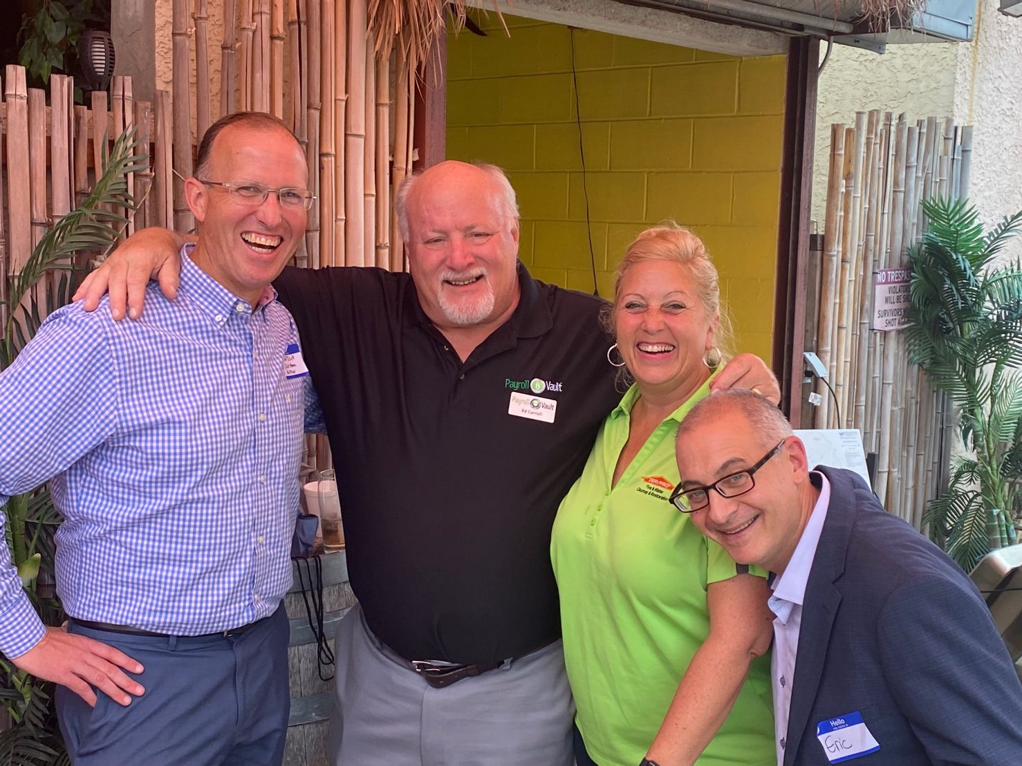 Four smiling adults wearing name tags embrace and laugh at an outdoor community gathering with bamboo décor.