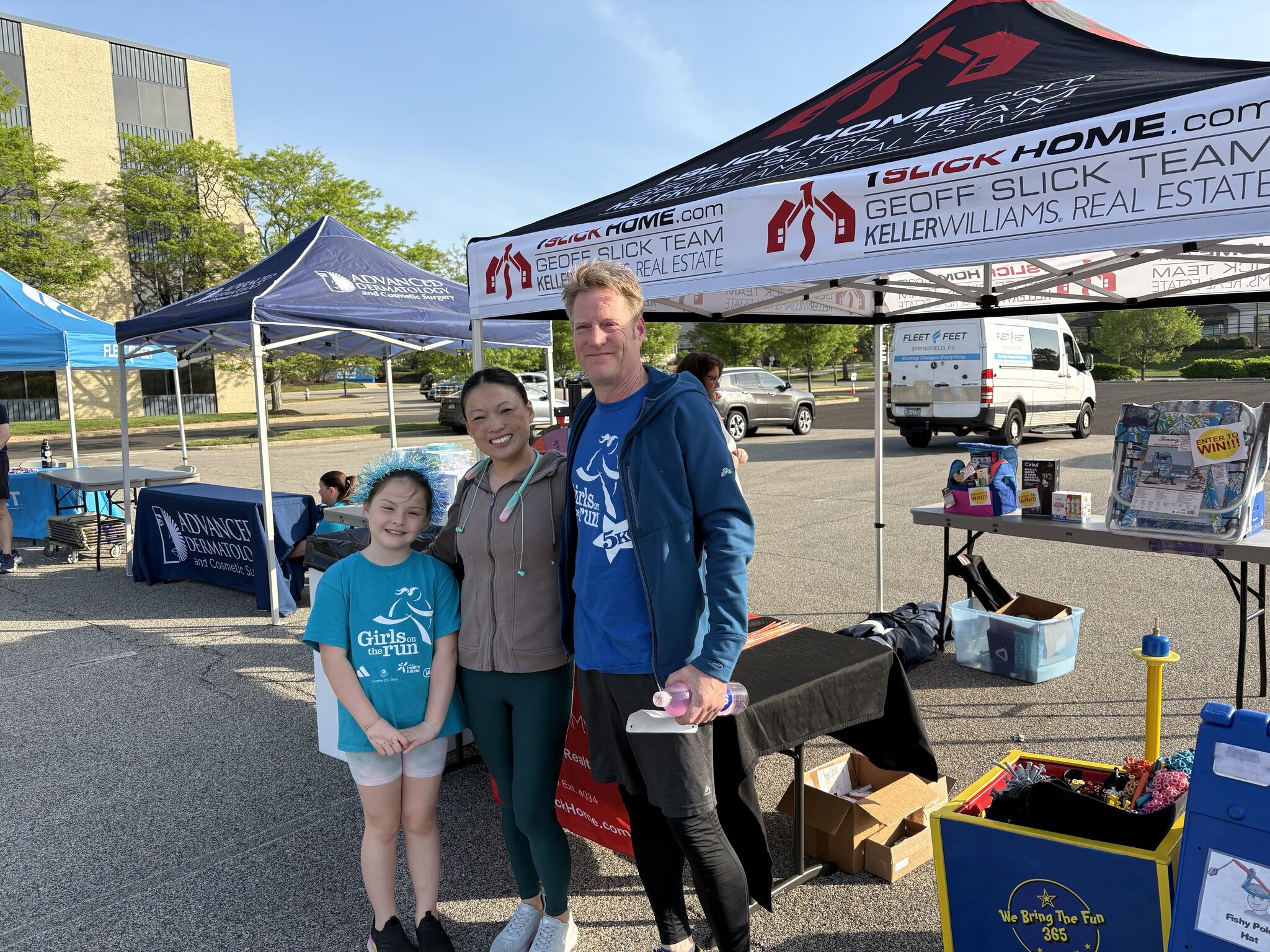 Man in blue jacket and two smiling women pose under a Keller Williams real estate tent at an outdoor community event.