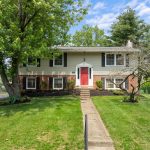 Split-level house with red front door, brick lower and beige siding upper, concrete path to the door and large trees in the yard.