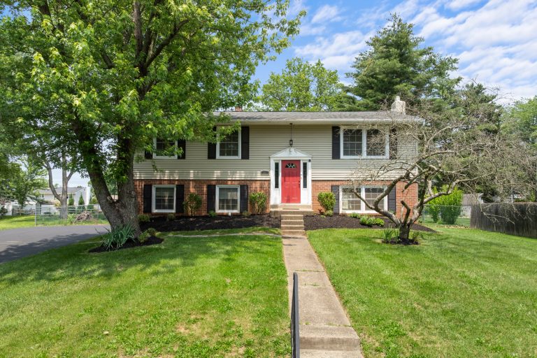 Split-level house with red front door, brick lower and beige siding upper, concrete path to the door and large trees in the yard.
