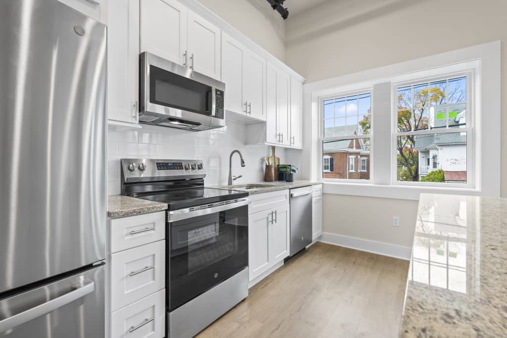 Bright kitchen at Lofts on Broad with white cabinets, stainless appliances, granite counters, and a large window facing brick homes.