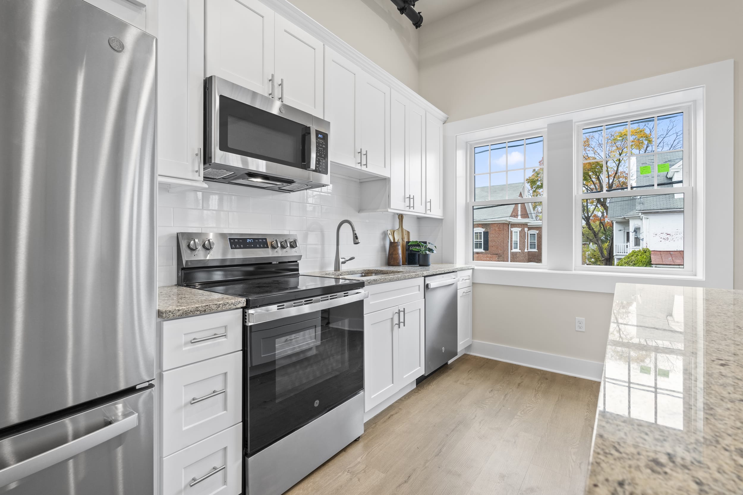 Bright kitchen at Lofts on Broad with white cabinets, stainless appliances, granite counters, and a large window facing brick homes.