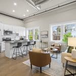 Open-plan loft with white kitchen, stainless fridge, island with black stools, tan leather chairs, and exposed ductwork