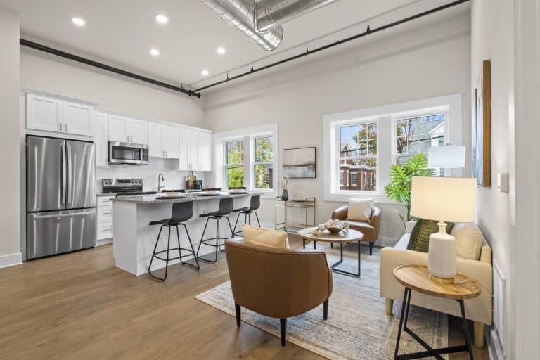 Open-plan loft with white kitchen, stainless fridge, island with black stools, tan leather chairs, and exposed ductwork