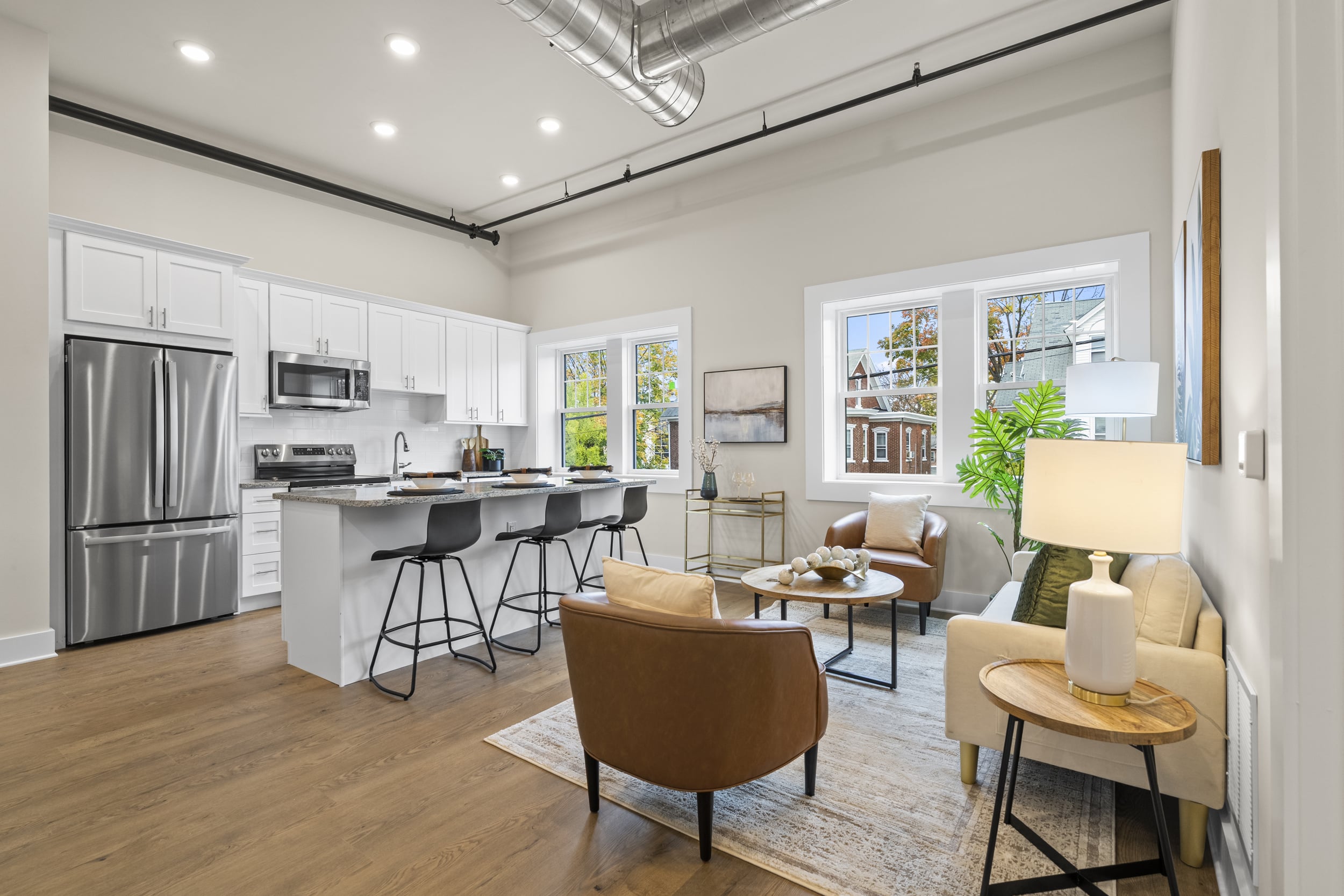 Open-plan loft with white kitchen, stainless fridge, island with black stools, tan leather chairs, and exposed ductwork