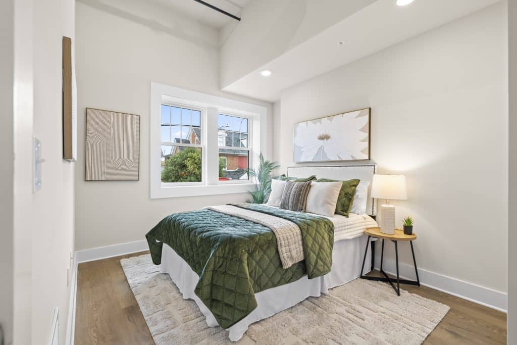 Bedroom at Lofts on Broad, Souderton, PA with a green quilted bed, white walls, window and round bedside table with lamp.