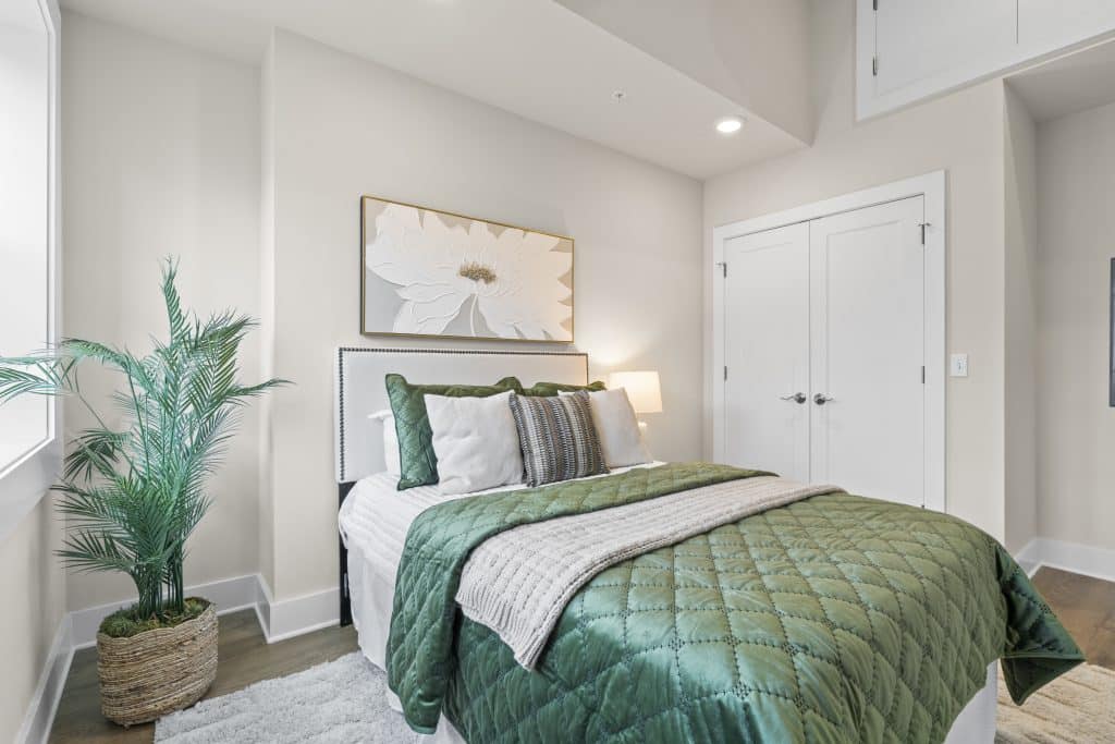 Lofts on Broad bedroom in Souderton with green quilted bed, white headboard, floral wall art, and potted palm.