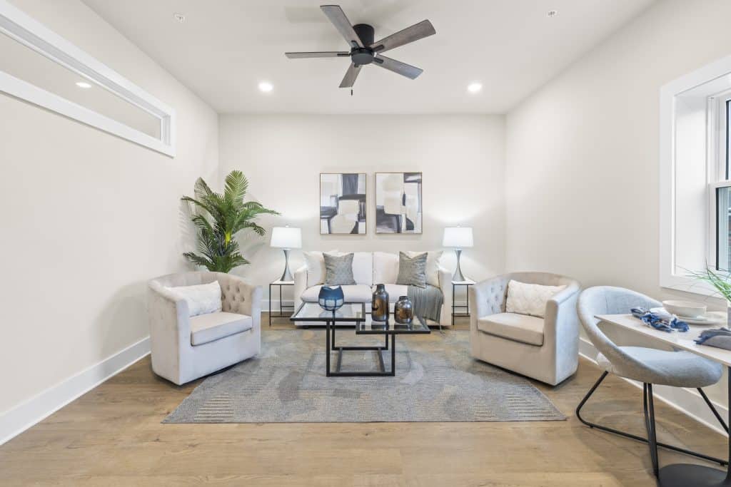 Modern living room with white sofa, two tufted armchairs, glass coffee table, ceiling fan, abstract art, and potted plant.