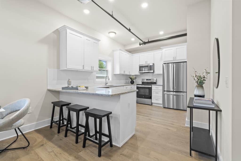 Bright kitchen at Lofts on Broad with white cabinets, granite island, three black stools, stainless appliances, and exposed black ceiling pipe.