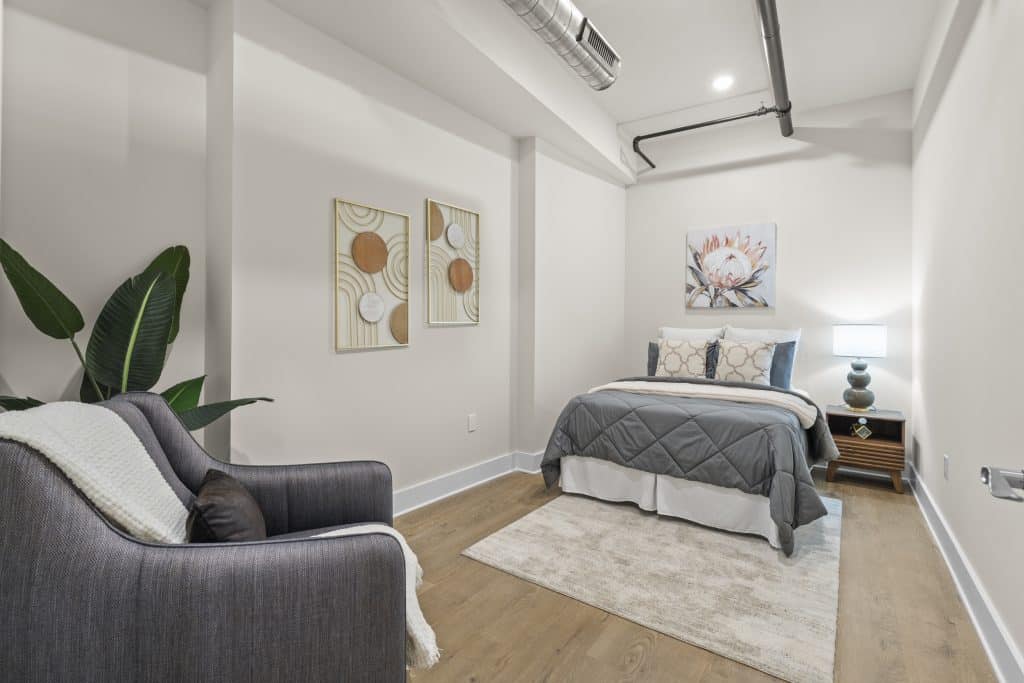 Small Lofts on Broad bedroom in Souderton, PA with gray bed, nightstand lamp, armchair, plant and exposed ductwork.