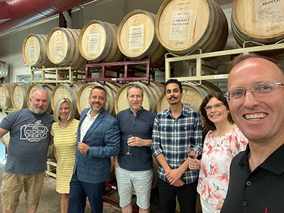 Seven people smiling and holding wine glasses in a winery barrel room during a 1SlickWine cabernet tasting.