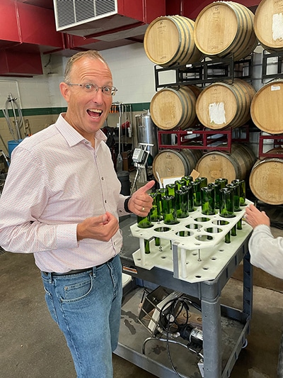 Smiling man giving thumbs-up beside a rack of green bottles for 1SlickWine Cabernet, with oak barrels stacked behind.