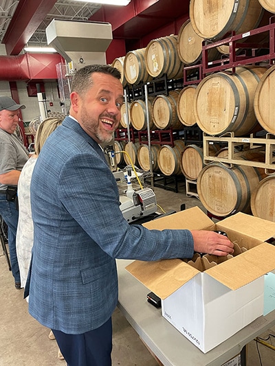 Man in blue plaid blazer smiling while packing 1SlickWine Cabernet bottles into a box in a barrel-lined winery.