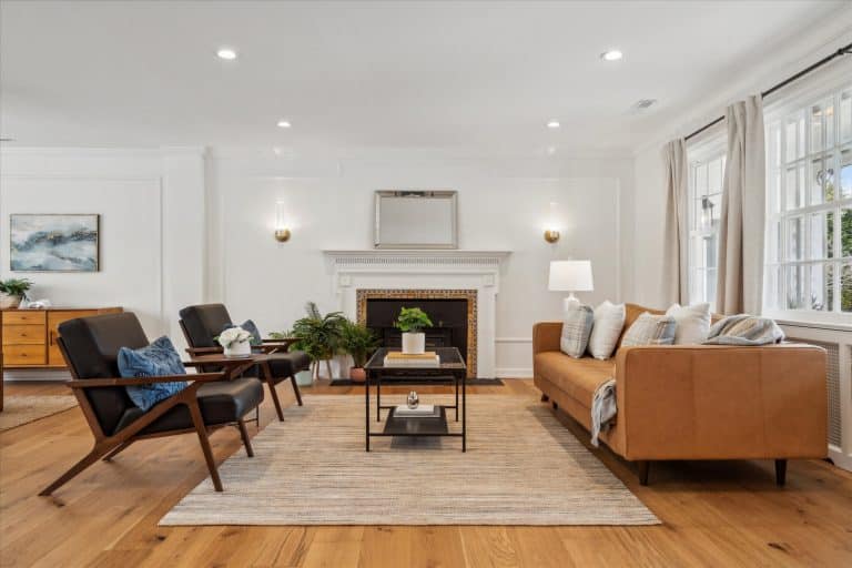 Bright living room with a tan leather sofa, two mid-century chairs, central coffee table, rug, and decorative fireplace