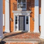 Front entrance of a red brick house with a dark gray paneled door framed by white pilasters, large fluted white columns, and brick steps.