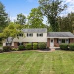 Split-level brick and siding suburban house with a manicured front lawn, shrubs, and trees against a wooded backdrop
