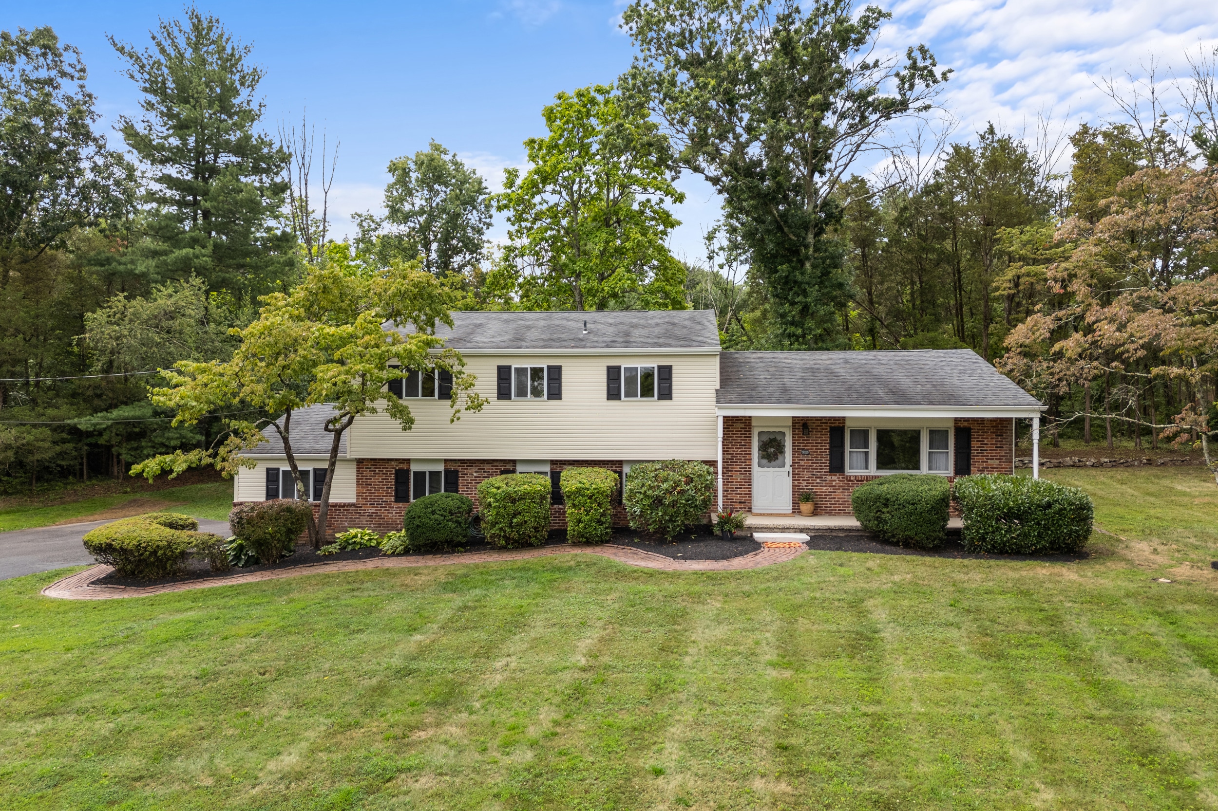 Split-level brick and siding suburban house with a manicured front lawn, shrubs, and trees against a wooded backdrop