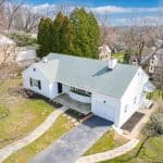 Single-story white house with a green roof, attached garage, curved front planter and stone walkway through a grassy yard, viewed from above.