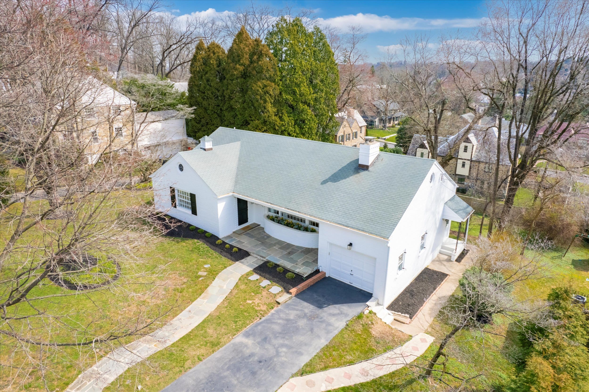 Single-story white house with a green roof, attached garage, curved front planter and stone walkway through a grassy yard, viewed from above.