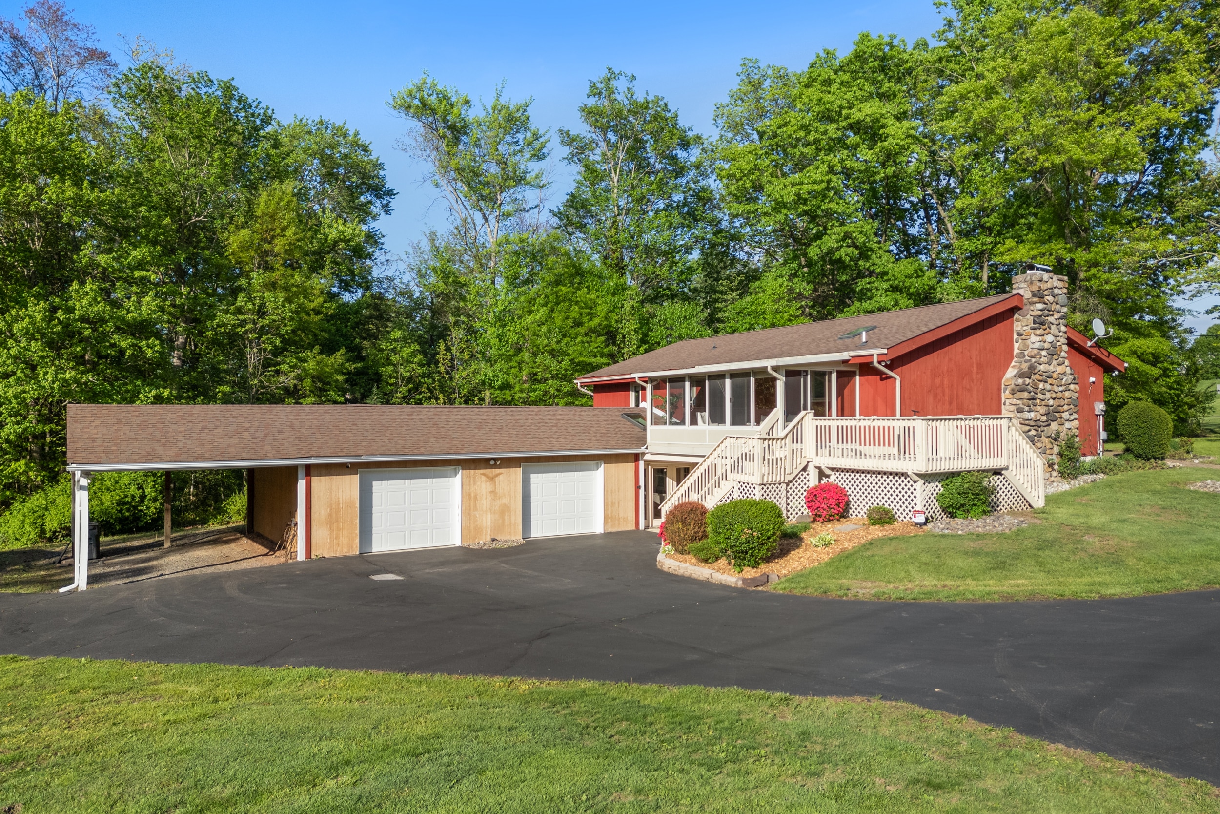 Split-level red house with a stone chimney, wraparound deck and three-car garage set against dense green trees and a paved driveway