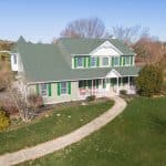 Two-story green-roof farmhouse with white porch, green shutters, curved walkway and large grassy yard in a rural setting.