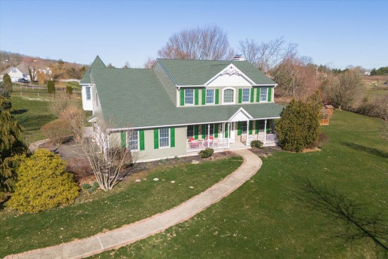 Two-story green-roof farmhouse with white porch, green shutters, curved walkway and large grassy yard in a rural setting.