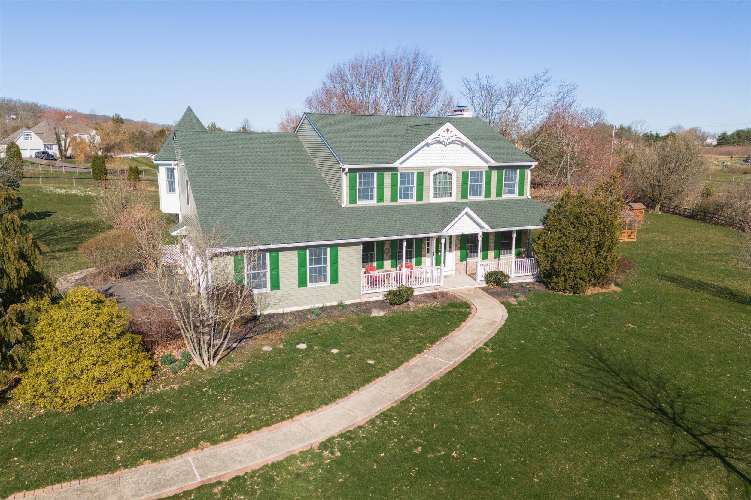 Two-story green-roof farmhouse with white porch, green shutters, curved walkway and large grassy yard in a rural setting.