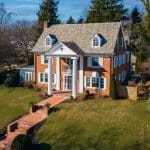Brick colonial house with white columns, dormer windows, slate roof, and a brick walkway across a sloped green lawn