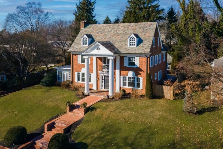 Brick colonial house with white columns, dormer windows, slate roof, and a brick walkway across a sloped green lawn