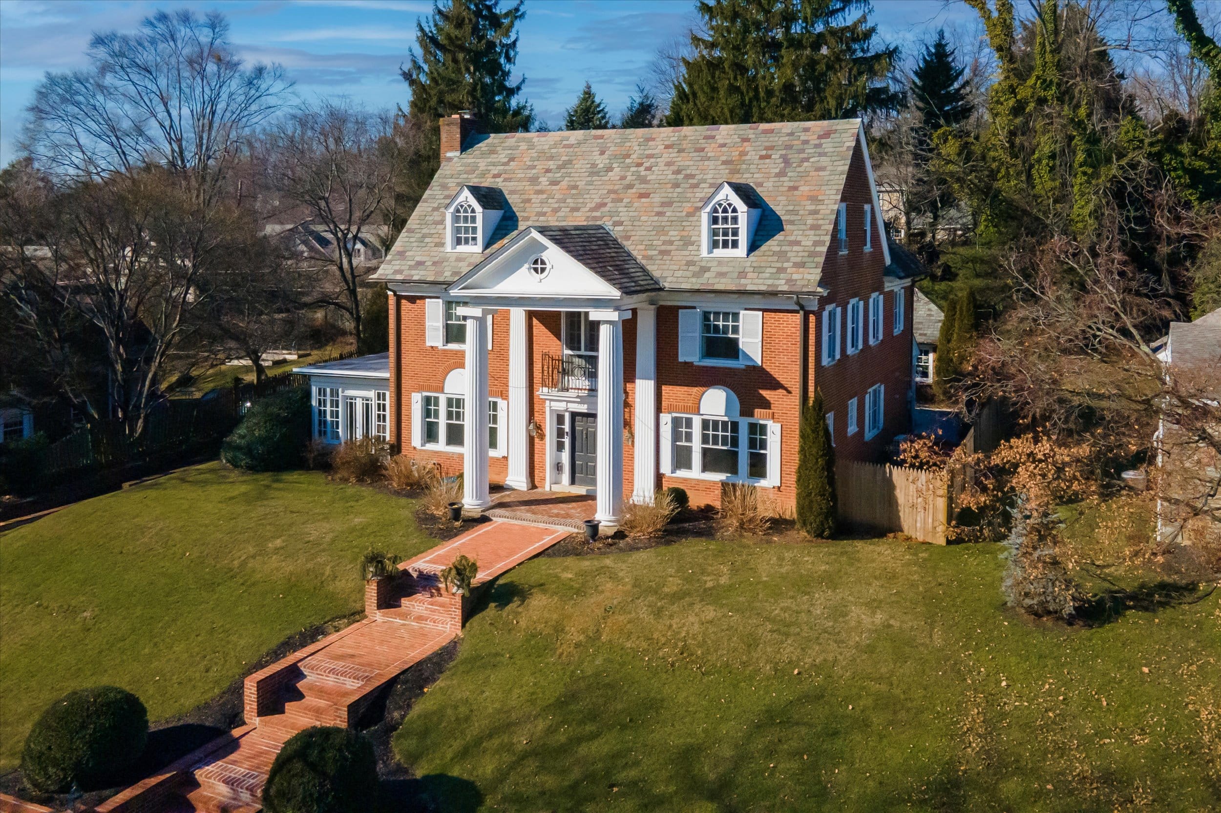 Brick colonial house with white columns, dormer windows, slate roof, and a brick walkway across a sloped green lawn